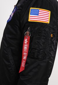 Černá bomber bunda se vyšitým záplatou americké vlajky, červeným štítkem "REMOVE BEFORE FLIGHT" a zipovými kapsami, s hladkým a lesklým povrchem.