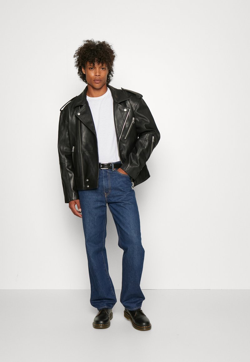 Giacca da biker in pelle nera con cerniere argentate, t-shirt bianca, jeans blue dritti e stivali neri. Il modello è in piedi contro uno sfondo semplice.