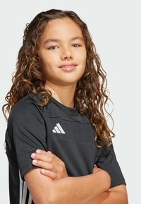 Camiseta atlética negra de manga corta, con un panel texturizado y tres franjas blancas a los lados. Modelo con cabello rizado sonriendo.