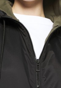 Mustatakki, jossa on irrotettava oliivinvihreä huppu, keskusvetoketju ja joustavat nauhat. Vaatteen päällä valkoinen paita.