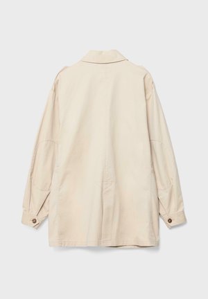 Beige pitkähiha takki, jossa on napitetut kalvosimet, kyynärpäiden paikkaukset ja rento malli, kuvattuna takaa yksiväristä taustaa vasten.