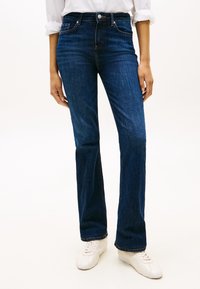 Personne portant un jean bootcut bleu foncé, des baskets blanches et une chemise blanche à manches longues avec les manches retroussées.