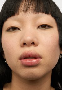 Nærbilde av et ansikt med rett svart hår, lys hud og perleøredobber. Glinsende lepper, subtile øyemakeup og naturlig hudtekstur synlig.