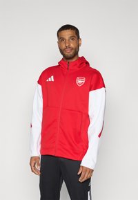 Red hooded jacket with white sleeves, featuring a front zipper, side pockets, and an Arsenal logo. Made from a smooth, structured fabric.