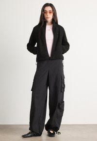 Svart fleecejakke med glidelås foran, rosa skjorte, oversized svarte cargo bukser med flere lommer, og svarte flate sko. Brune solbriller.