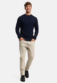 Marineblå kabelstrikket genser med rund hals, kombinert med lysebeige bukser og svarte ankelboots, på en modell som står avslappet.