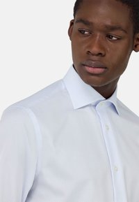 Camicia azzurro chiaro a maniche lunghe realizzata in tessuto a quadretti. Presenta un colletto con bottoni e una sottile texture con piccoli quadrati.
