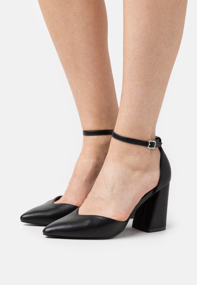 Pieds d'une femme portant des chaussures à talons blocs en cuir noir à bout pointu avec brides autour de la cheville, sur fond blanc.