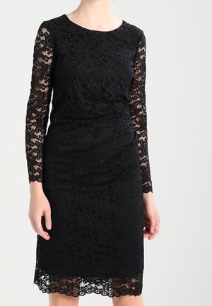 Femme portant une robe noire en dentelle florale longueur genou, avec de longues manches en dentelle transparente et un col rond.