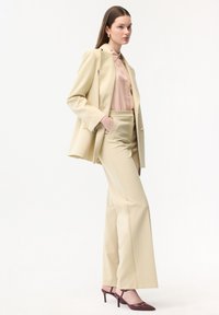 Mujer con traje beige y blusa rosa pálido, calzando zapatos de tacón burdeos, de pie con una mano en el bolsillo sobre un fondo blanco.