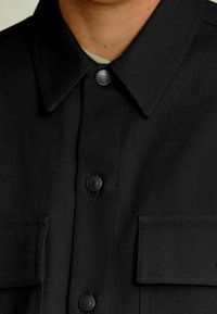 Sort skjorte med spidst krave, synlige knapper og forlommer. Tekstureret stof med et diagonalt mønster.