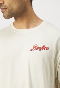 Kreminbaltas kokvilnas T-krekls ar sarkanu izšūtu "Benfica" logo kreisajā krūtī, ar apaļu kaklu un īsām piedurknēm.