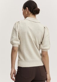 Beige Strickpullover mit einem Kragen, Puffärmeln und geripptem Saum. Weiche Textur mit subtilen Farbvariationen. Kombiniert mit dunkelbraunen Hosen.