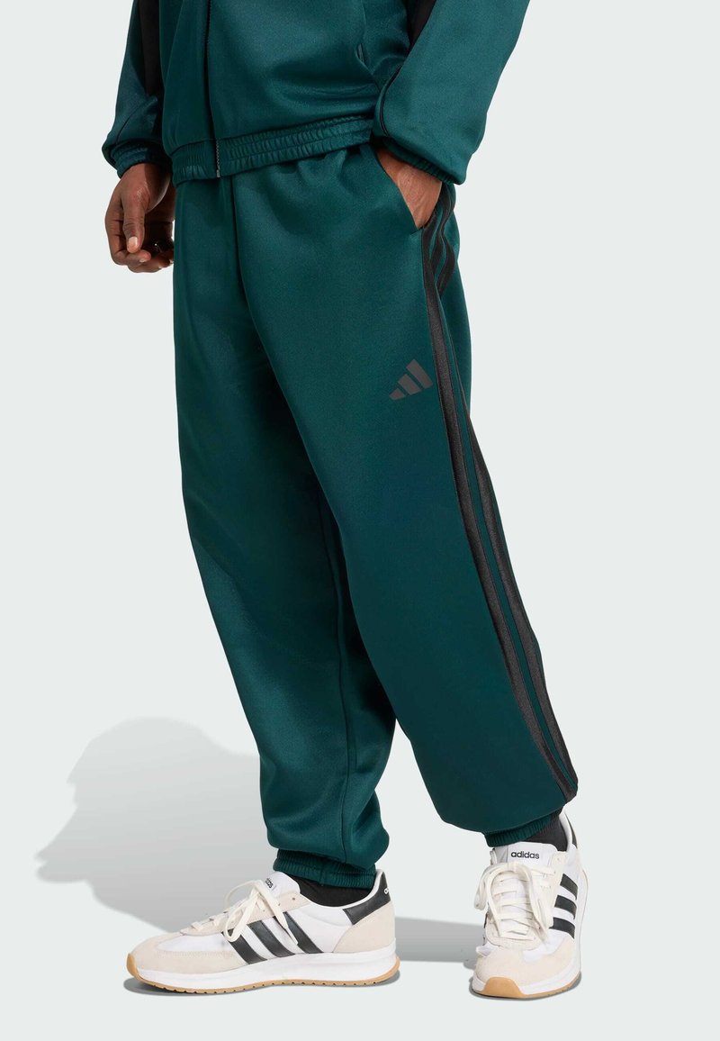 Pantalons de survêtement verts en matériau lisse avec une taille élastique, des poches latérales et des rayures noires. Associés à des baskets blanches avec des accents noirs.