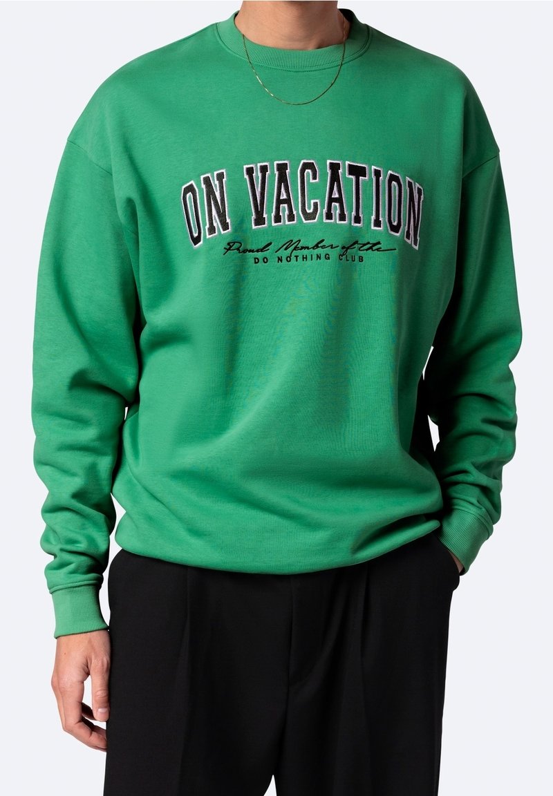 Personne portant un sweat-shirt vert avec le texte « EN VACANCES Membre fier du CLUB DU NE RIEN FAIRE », associé à un pantalon noir.