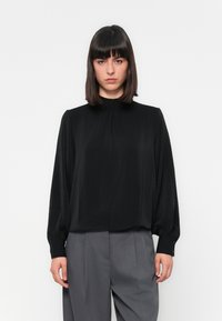 Donna con capelli scuri e lisci, che indossa una blusa nera a maniche lunghe e pantaloni grigi a vita alta su uno sfondo bianco.