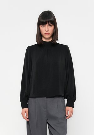 Woman with straight dark hair wearing a black long-sleeve blouse and gray high-waisted trousers against a white background.