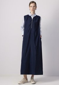 Vestido sin mangas de color azul marino con cremallera frontal, bolsillos laterales y un ajuste relajado. Llevado sobre una camisa de cuello azul claro. Tejido texturizado.