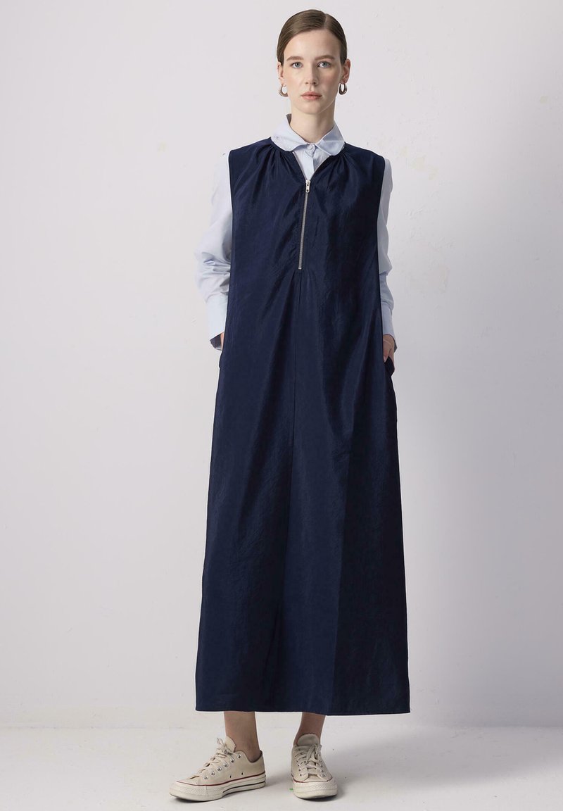 Vestido sin mangas de color azul marino con cremallera frontal, bolsillos laterales y un ajuste relajado. Llevado sobre una camisa de cuello azul claro. Tejido texturizado.