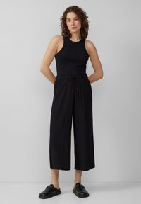 Zwarte mouwloze top met kleine versieringen, gecombineerd met losse zwarte cropped broeken. Heeft een elastische taille en een gestrikte bovenkant. Zwart schoeisel.
