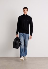 Fekete cipzáros pulóver, világoskék farmernadrág, fekete bőr táska. Hétköznapi cipő és minimalista design, egyszínű háttér előtt.