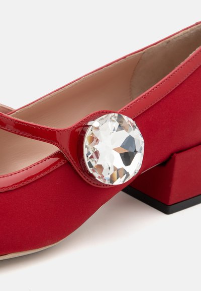 Piros lakkbőr cipő, lekerekített orral, lapos sarokkal, és egy nagy, átlátszó, csiszolt drágakő díszítéssel az oldalon. Simaság jellemzi, fényes felülettel.