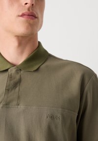 Olivgrünes Poloshirt mit strukturierter Oberfläche, geripptem Kragen und subtiler Logo-Stickerei. Knopfverschlussdetails auf der Vorderseite.