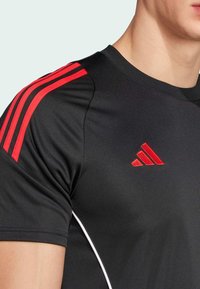 Nära bild av en person iförd en svart sporttröja med röda ränder på axeln och en röd Adidas-logga på bröstet.