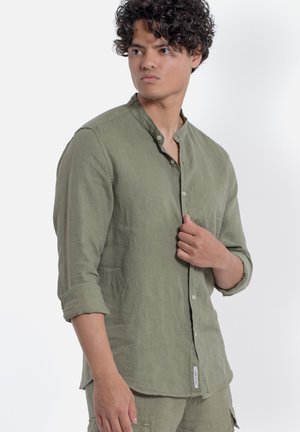 Giovane uomo con capelli ricci che indossa una camicia di lino verde oliva con bottoni e maniche arrotolate, guardando di lato su uno sfondo bianco.