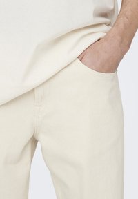 Light beige cotton trousers with a straight fit, featuring a side pocket and visible stitching along the seams. Smooth texture, minimal design.