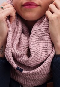 Knitted infinity scarf in soft, light pink yarn. Features a textured rib pattern and a small navy label tag, worn loosely around the neck.