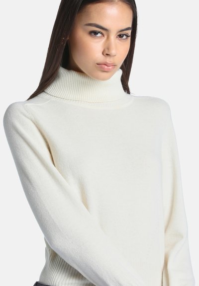 Jeune femme aux longs cheveux foncés portant un pull à col roulé blanc sur un fond clair uni.