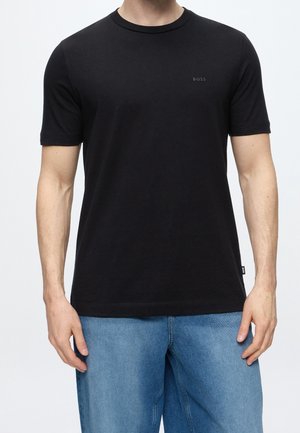 Homme portant un t-shirt noir uni à manches courtes avec le logo "BOSS" sur la poitrine et une étiquette latérale, associé à un jean en denim bleu clair.