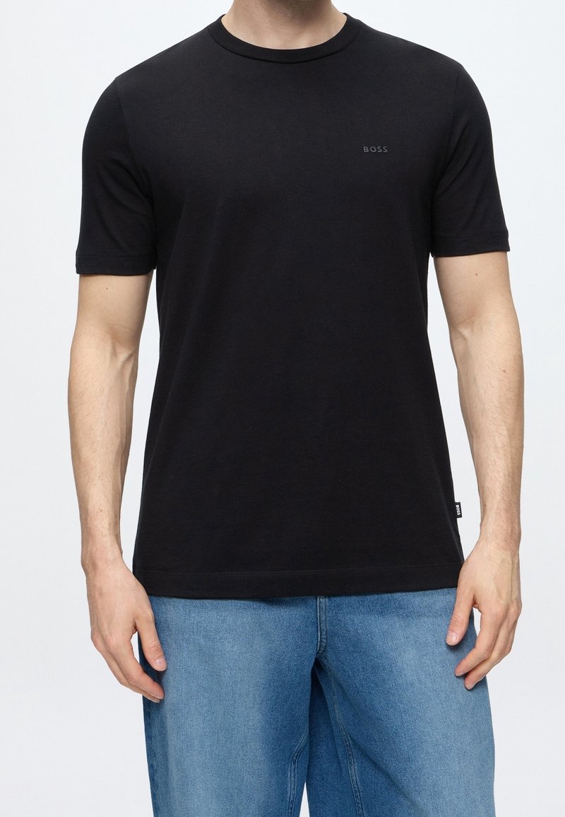 Homme portant un t-shirt noir uni à manches courtes avec le logo "BOSS" sur la poitrine et une étiquette latérale, associé à un jean en denim bleu clair.