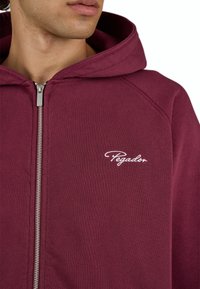 Male wearing a maroon zip-up hoodie with a hood and white "Pegador" logo on the left chest against a white background.
