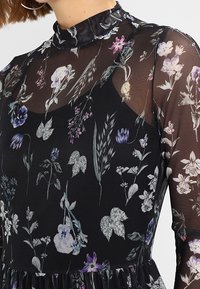 Robe en tissu noir transparent avec un motif floral violet, blanc et vert. Elle présente un col montant et des manches longues bouffantes. Texture délicate.
