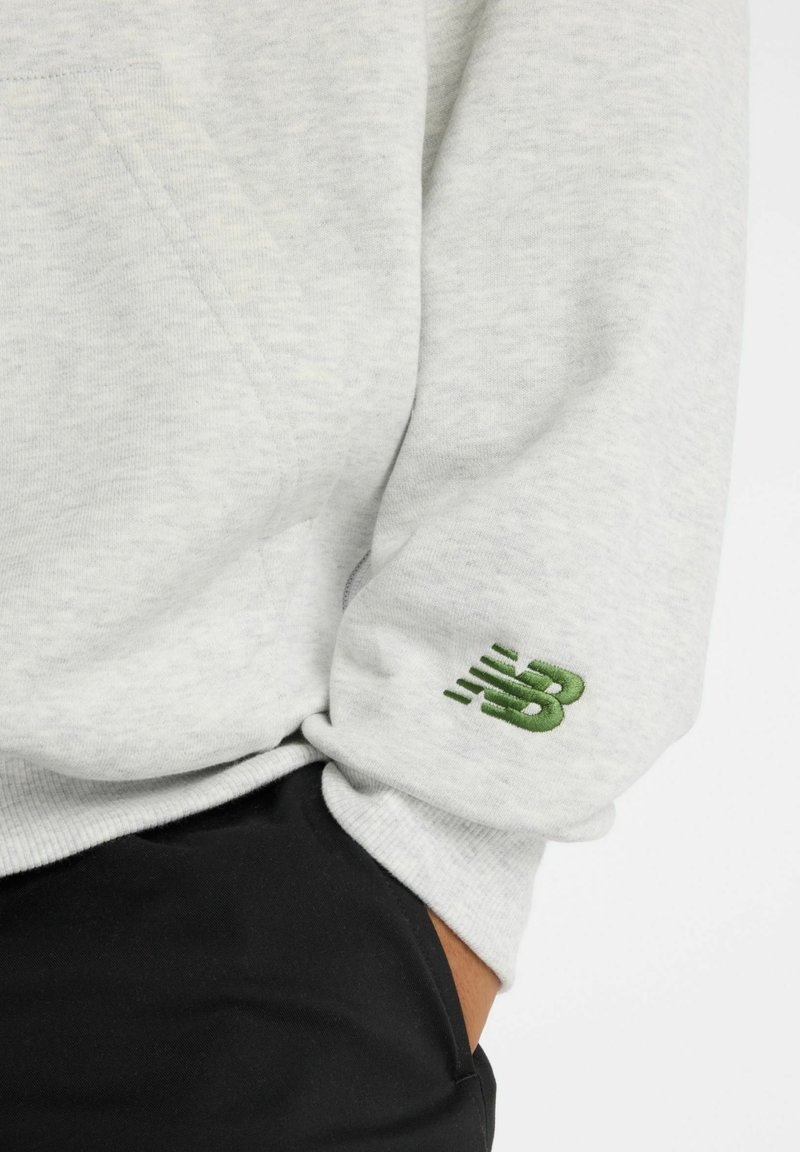 Helles graues Sweatshirt mit einer weichen Textur; ausgestattet mit einem grünen gestickten Logo auf dem Ärmel und einer geräumigen Tasche auf der Vorderseite.