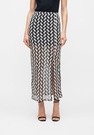 Jupe longue à motif zigzag noir et blanc avec une fente devant, portée avec un haut noir et des talons noirs à bride cheville.