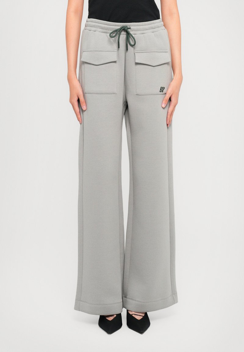 Light gray wide-leg sweatpants in a soft fabric, featuring two front pockets, an elastic waistband with a green drawstring, and logo detail.