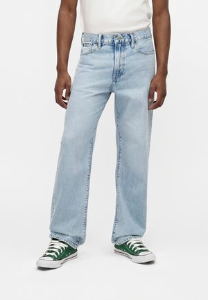 Személy világoskék, bő fazonú farmernadrágot, fehér felsőt és zöld alacsony szárú tornacipőt visel fehér cipőfűzővel, sima háttér előtt áll.