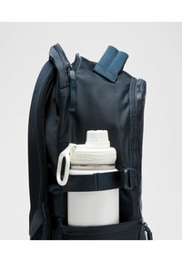 Tamsiai mėlynas kuprinė su užtrauktuko uždarymu, kuriame yra balta vandens butelis, laikomas dokumentu šoniniame kišenėje. Tekstūruota medžiaga.