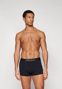 Izmos férfi fekete ellesse boxeralsót visel, egy sima szürke háttér előtt áll, és balra néz.