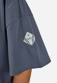 Μανίκι σκούρου μπλε πουκαμίσου με λευκό σημείο που φέρει γεωμετρικό λογότυπο και την επιγραφή "PEGADOR" σε ανοιχτού τόνου δέρματος χέρι.