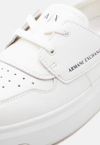 Λευκό δερμάτινο αθλητικό παπούτσι με λευκά κορδόνια, διάτρητη μύτη και τη σήμανση "ARMANI EXCHANGE" στο πλάι καθώς και "A|X" στη γλώσσα.