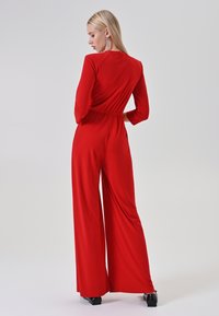 Donna bionda con orecchini lunghi che indossa un'intera tuta rossa a maniche lunghe e scarpe nere, in piedi e girata di spalle su uno sfondo semplice.