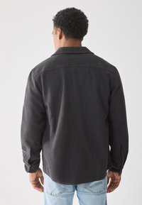 Camisa de un gris oscuro y texturizado con cuello de punta, mangas largas y puños abotonados, combinada con jeans azul claros. Se muestra la vista trasera.