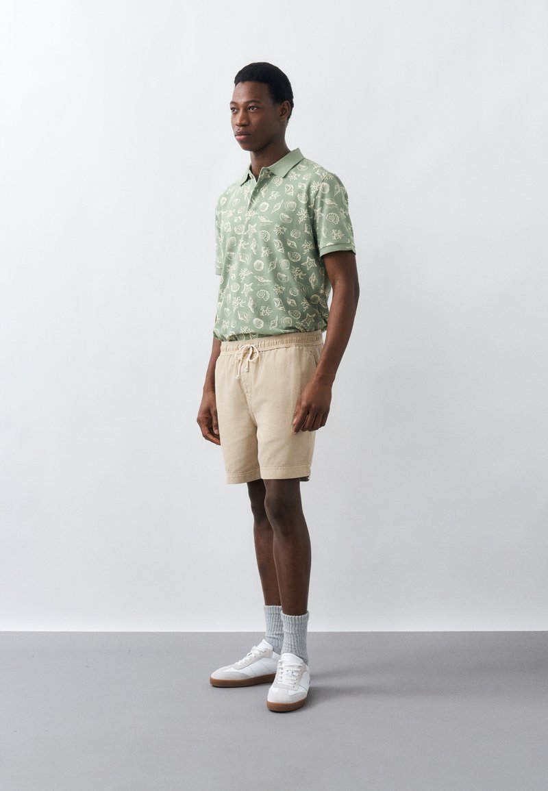 Giovane uomo in piedi che indossa una polo verde chiaro stampata, pantaloncini beige con coulisse, calzini grigi e scarpe da ginnastica bianche su sfondo bianco.