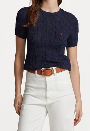 Marineblå kortærmet strikket sweater med kabelmønster, med en lomme og et lille rødt emblem. Kombineret med hvide højtaljede bukser og et tanfarvet bælte.