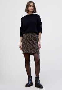 Suéter de punto negro con detalles de botones en el hombro, combinado con una falda marrón con estampado de leopardo. La modelo lleva botas negras hasta el tobillo y medias transparentes.