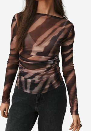 Vrouw draagt een doorzichtig lang mouwen top met een bruin-zwart tie-dye patroon, gecombineerd met zwarte high-waisted jeans.